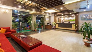 A hotel lobby with a reception desk, red sofas, a glass coffee table, and potted plants. - Hotel Queens Park (Pokhara)