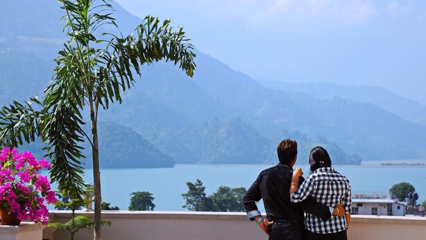 A couple standing on a balcony overlooking a lake with mountains in the background. - Hotel Queens Park (Pokhara)