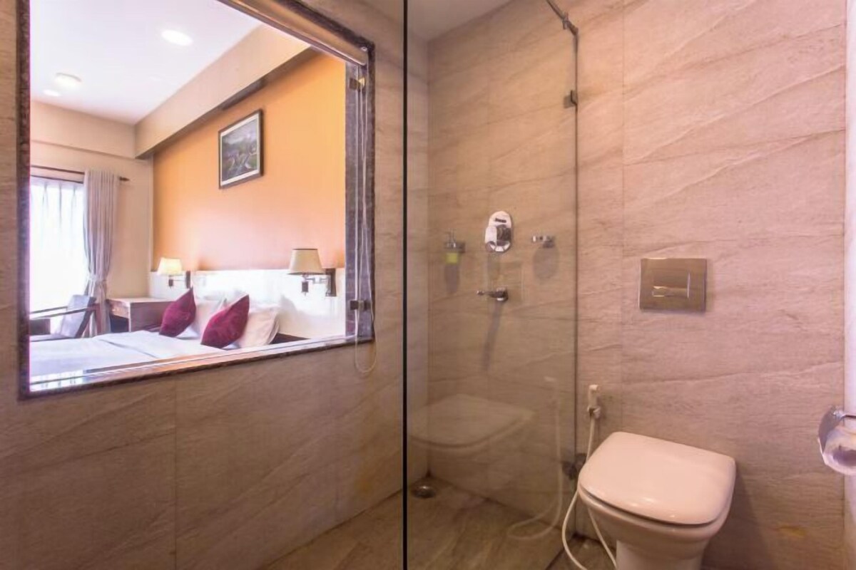 A hotel bathroom with a glass-enclosed shower, a toilet, and a bed visible through a window.