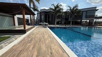 Un espace piscine moderne avec une terrasse en bois, un coin salon couvert et une voiture blanche garée près de la piscine.