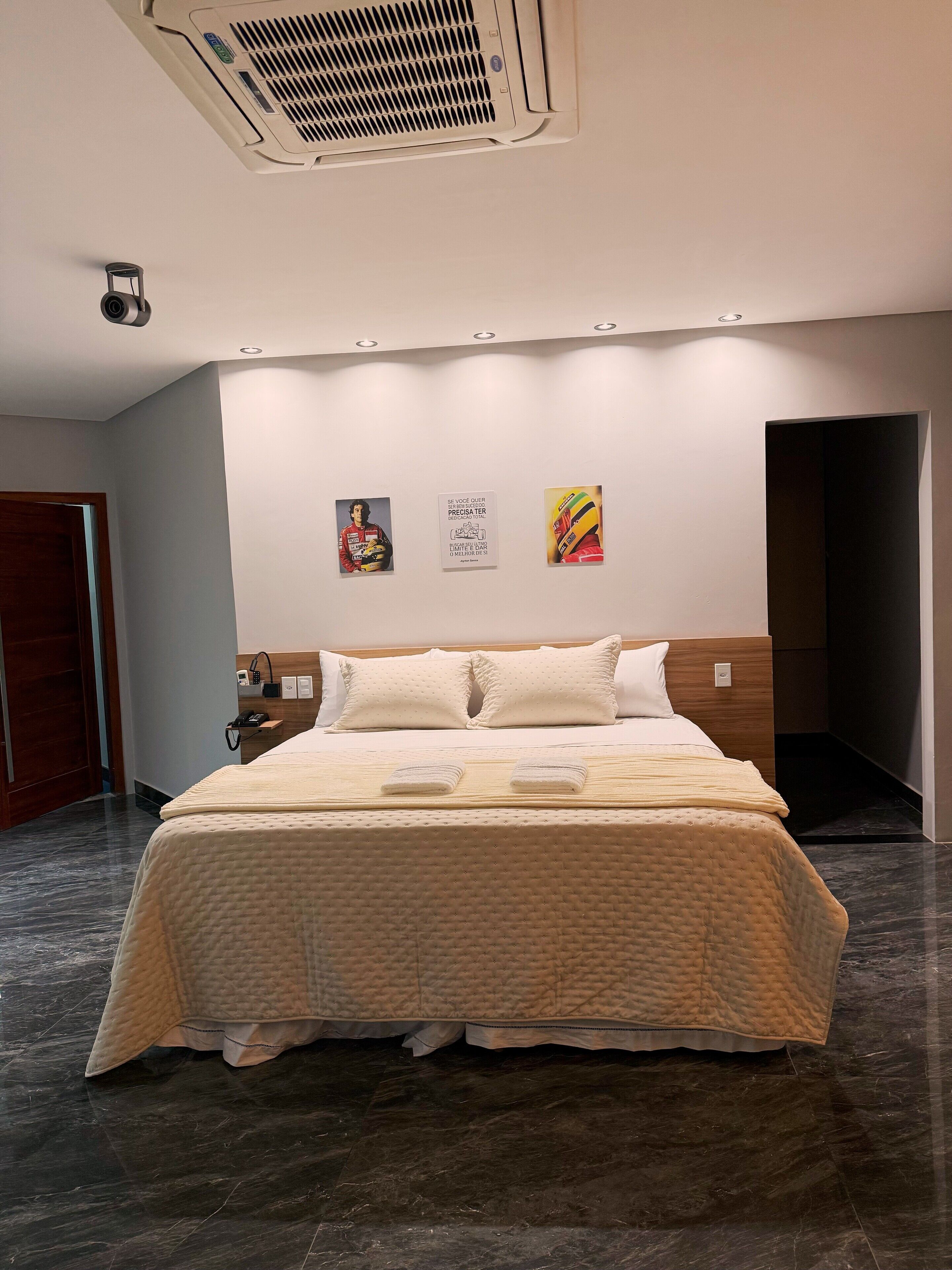 A modern hotel room with a large bed, a wall-mounted air conditioner, and three framed pictures.