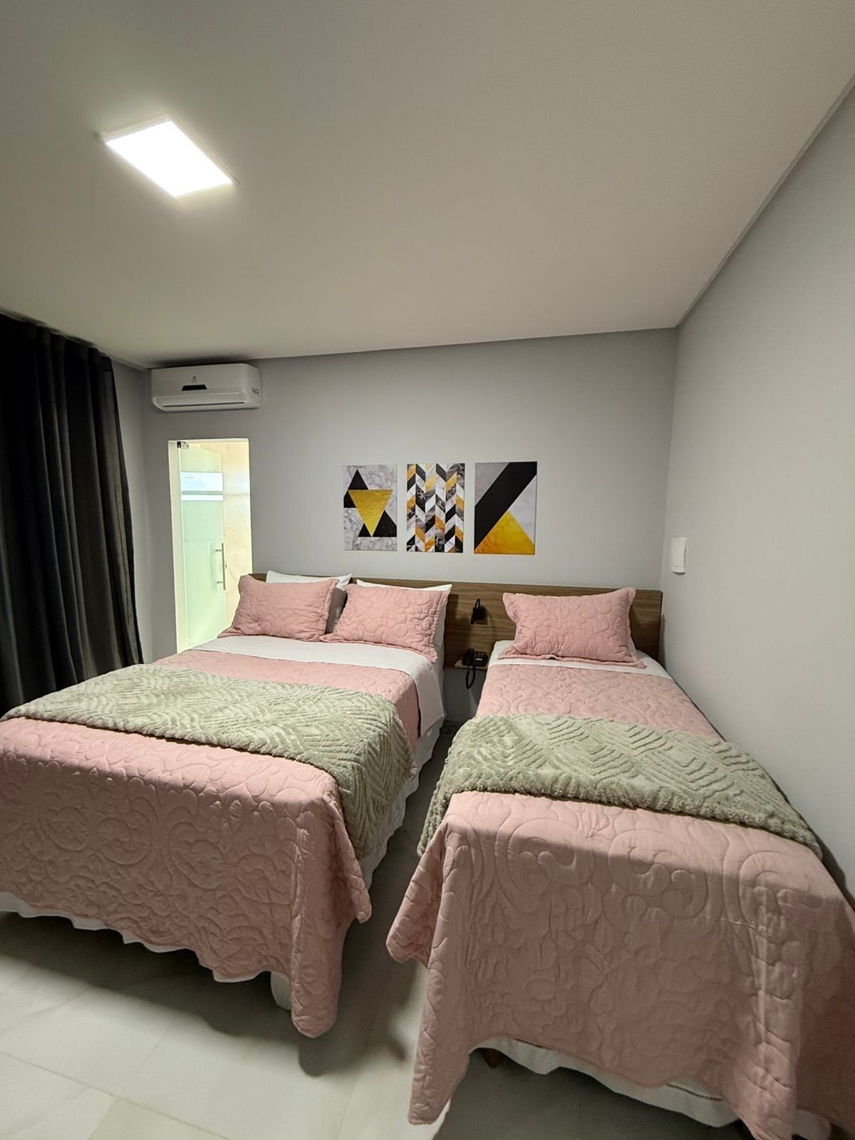 Habitación de hotel con dos camas, una lámpara de techo, un equipo de aire acondicionado y tres obras de arte en la pared.