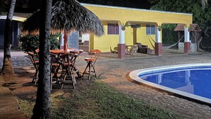 A poolside area with a yellow building, outdoor seating, and palm trees.