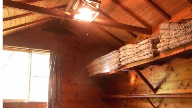 A wooden cabin interior with a sloped ceiling, a window with curtains, and a stack of blankets on a shelf. - Gujohachimanshizenen - Bungalow (Gujo)