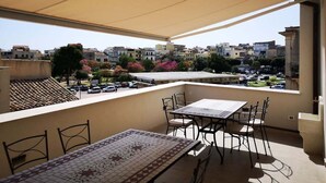A balcony with a table and chairs, a view of buildings, and a parked car in the background.