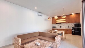 A modern living room with a sectional sofa, a coffee table, and a dining area with a table and chairs. The kitchen features wooden cabinets, a black refrigerator, and a marble backsplash.