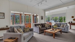 Amplia sala de estar con dos sofás, una mesa de centro y una mesita auxiliar de madera. Grandes ventanales ofrecen luz natural y vistas al verdor exterior.