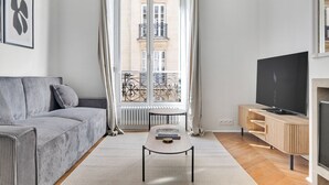 Ein modernes Wohnzimmer mit einem grauen Sofa, einem Fernseher auf einem Holzboden, einem kleinen Tisch und Blick auf ein Gebäude durch ein großes Fenster.