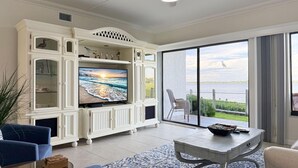A spacious living room with a large flat-screen TV, white cabinetry, a ceiling fan, and a view of the water through large sliding glass doors. - Tamarind – 2BR Condo with Lemon Bay Views (Englewood)