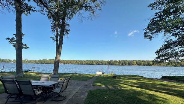 Área externa com mesas e cadeiras, um caminho pavimentado e vista para um lago com árvores ao fundo.
