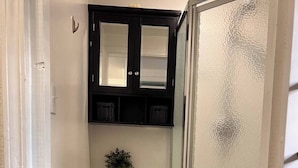 A compact bathroom with a toilet, a small shelf above the toilet, a potted plant, and a shower area with a glass door. - Cozy Suburb Studio (Everett)