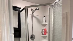 A corner shower with a glass door, a white toilet, and a shelf with toiletries. - Cozy Suburb Studio (Everett)