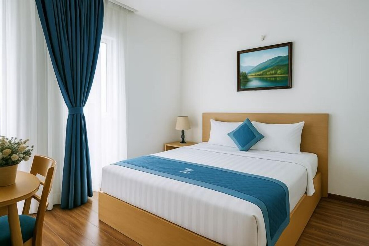 A hotel room with a bed, a nightstand, a lamp, a painting on the wall, and a window with curtains.