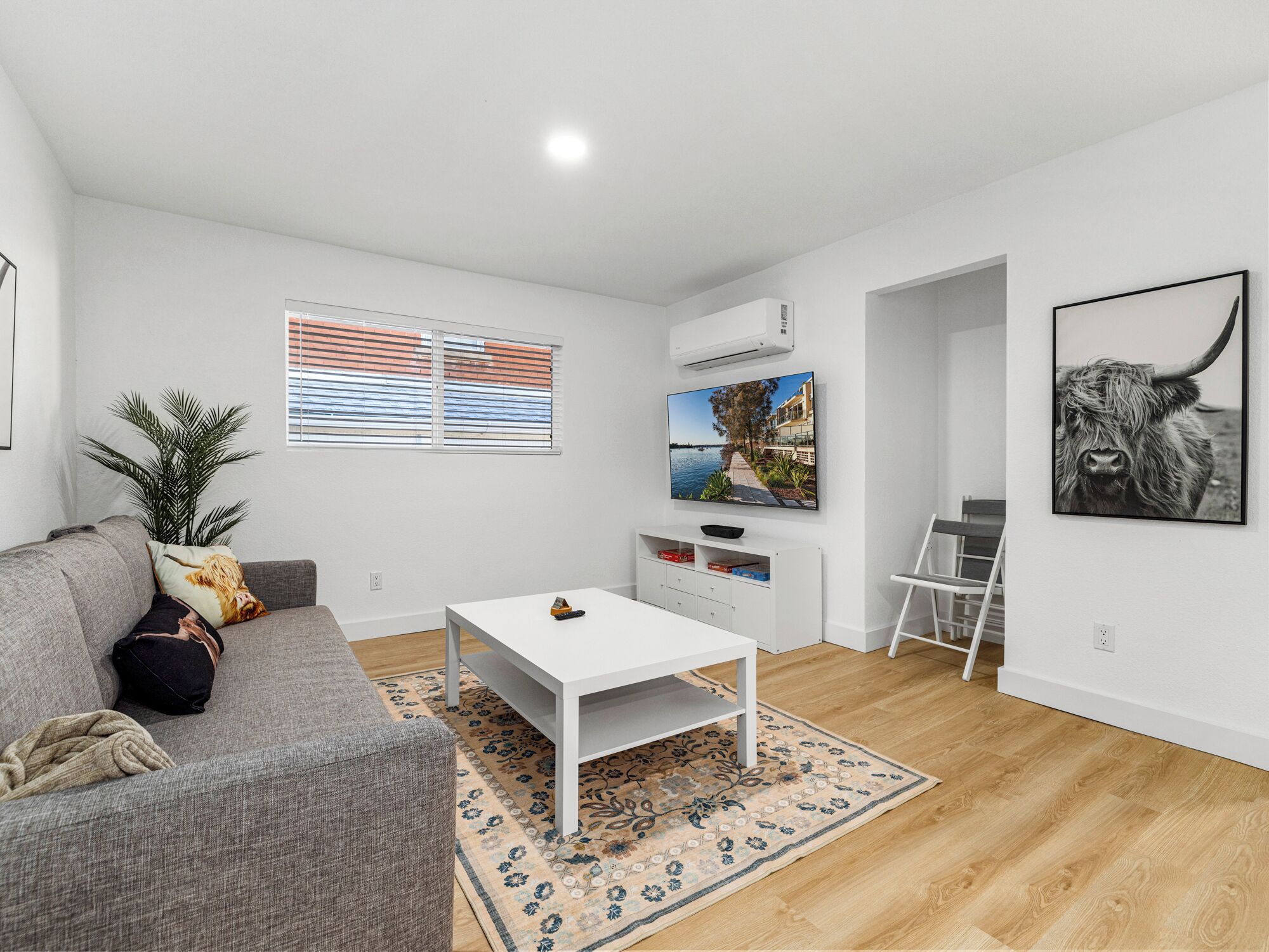 Un salon moderne avec un canapé gris, une table basse blanche, un téléviseur fixé au mur et une grande fenêtre avec des stores.