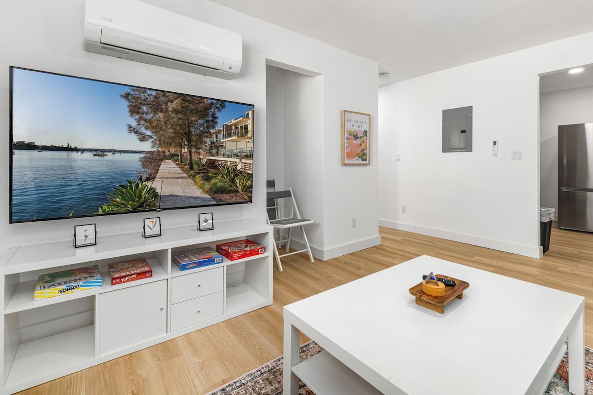 Un salon moderne doté d’un grand téléviseur à écran plat fixé au mur, d’un meuble de rangement blanc contenant des livres et des jeux, d’une table basse blanche et offrant une vue sur un plan d’eau par la fenêtre.