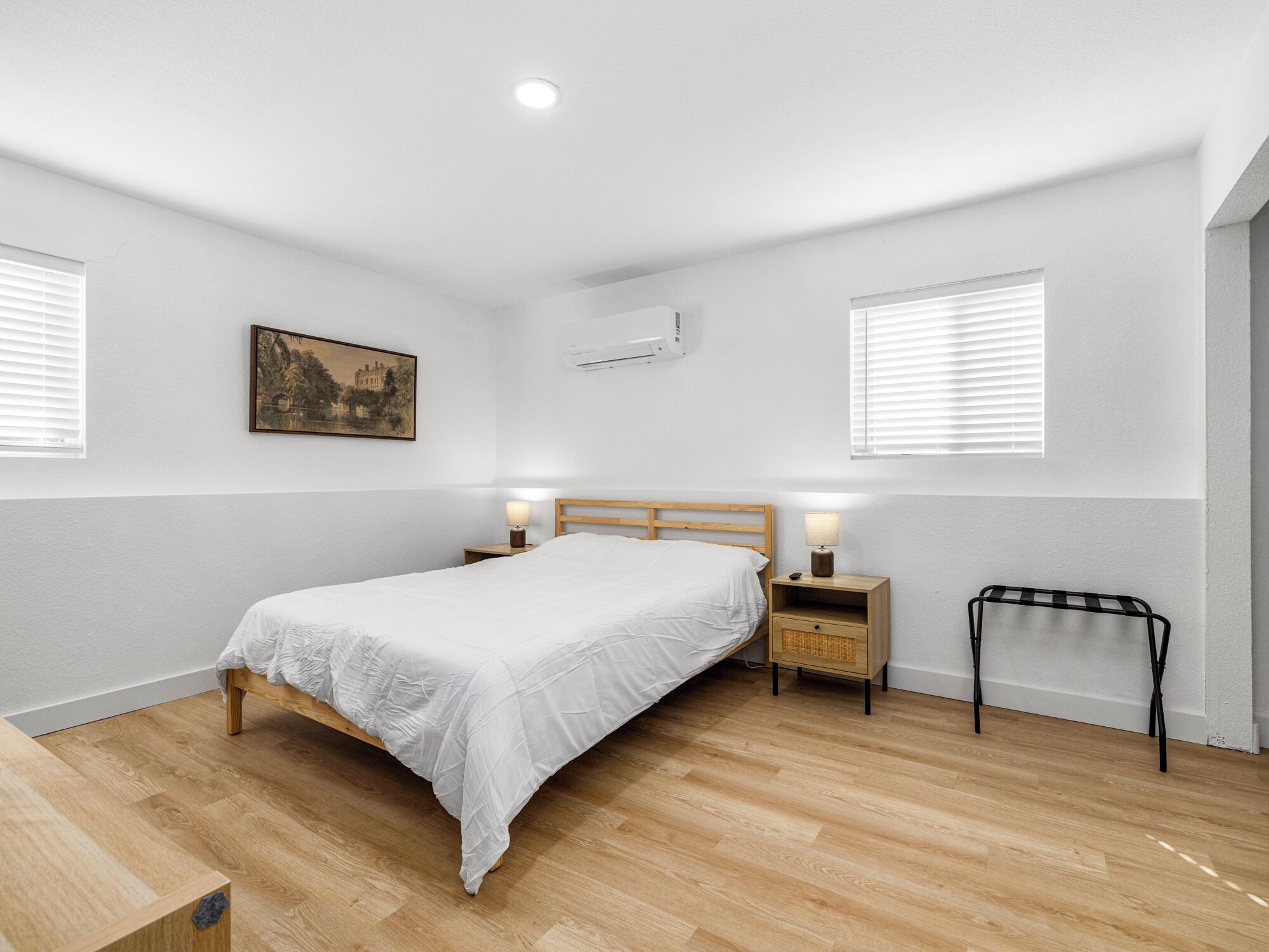 A bedroom with a wooden bed, bedside tables with lamps, a wall-mounted air conditioner, and a painting on the wall.