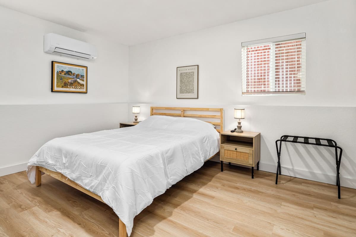 Une chambre avec un lit en bois, deux tables de chevet avec des lampes, une fenêtre avec des stores, un climatiseur et deux tableaux encadrés accrochés au mur.