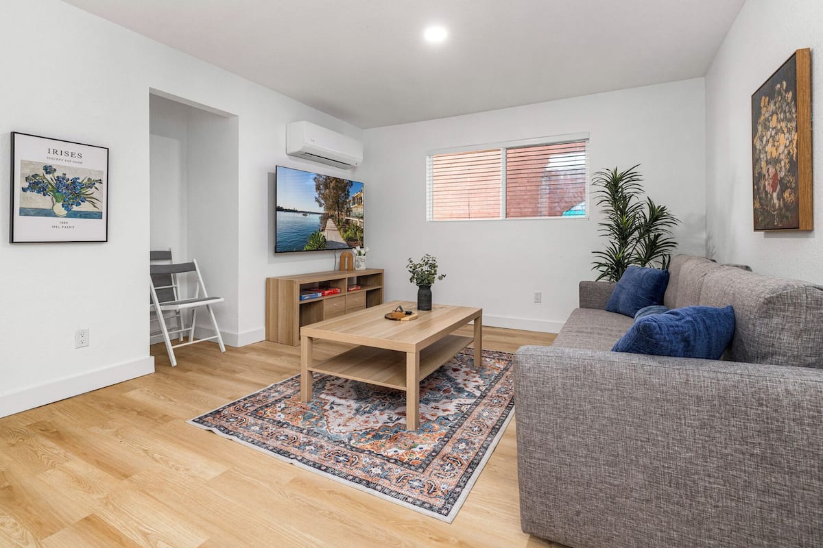 Un salon moderne avec un canapé gris, une table basse en bois, un téléviseur fixé au mur et un tapis à motifs sur le sol.