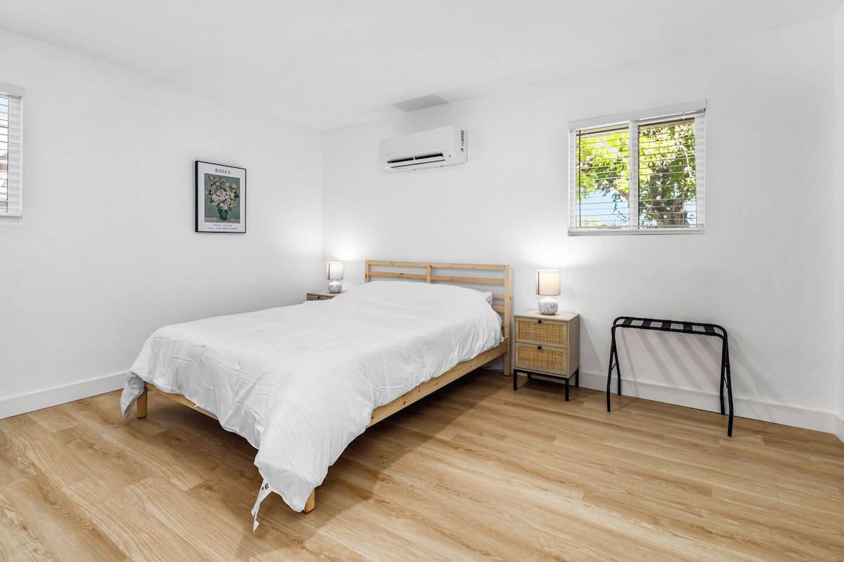 Une chambre avec un lit en bois, du linge de lit blanc, une table de chevet avec une lampe, un tableau au mur et une fenêtre avec des stores.
