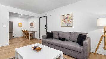 Een moderne woonkamer met een grijze bank, een witte salontafel en een eethoek met houten stoelen.