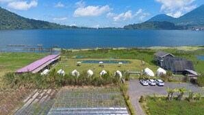 A lakeside property with a greenhouse, a covered outdoor seating area, and a parking lot with cars. - Butterfly Bali Glamping (Baturiti)