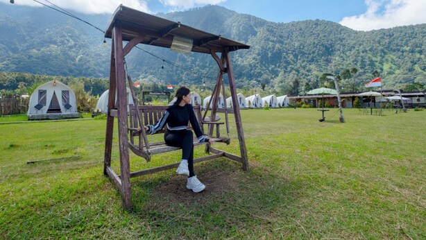 A person sitting on a wooden swing in a grassy area with tents and a mountain in the background. - Butterfly Bali Glamping (Baturiti)
