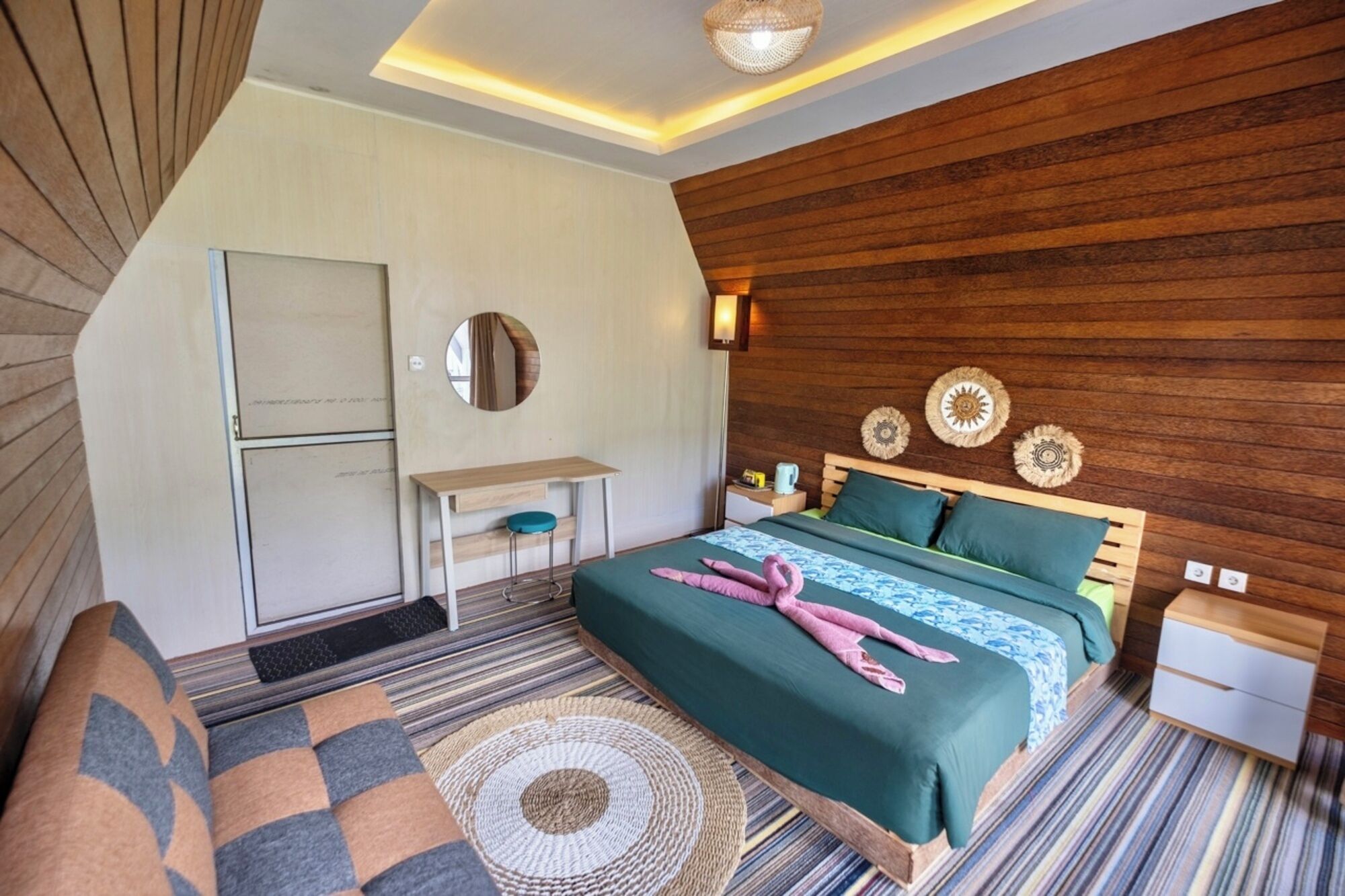 Una habitación de hotel moderna con una pared de acento de madera, una cama con ropa de cama verde, un escritorio con una silla y un espejo redondo pequeño.