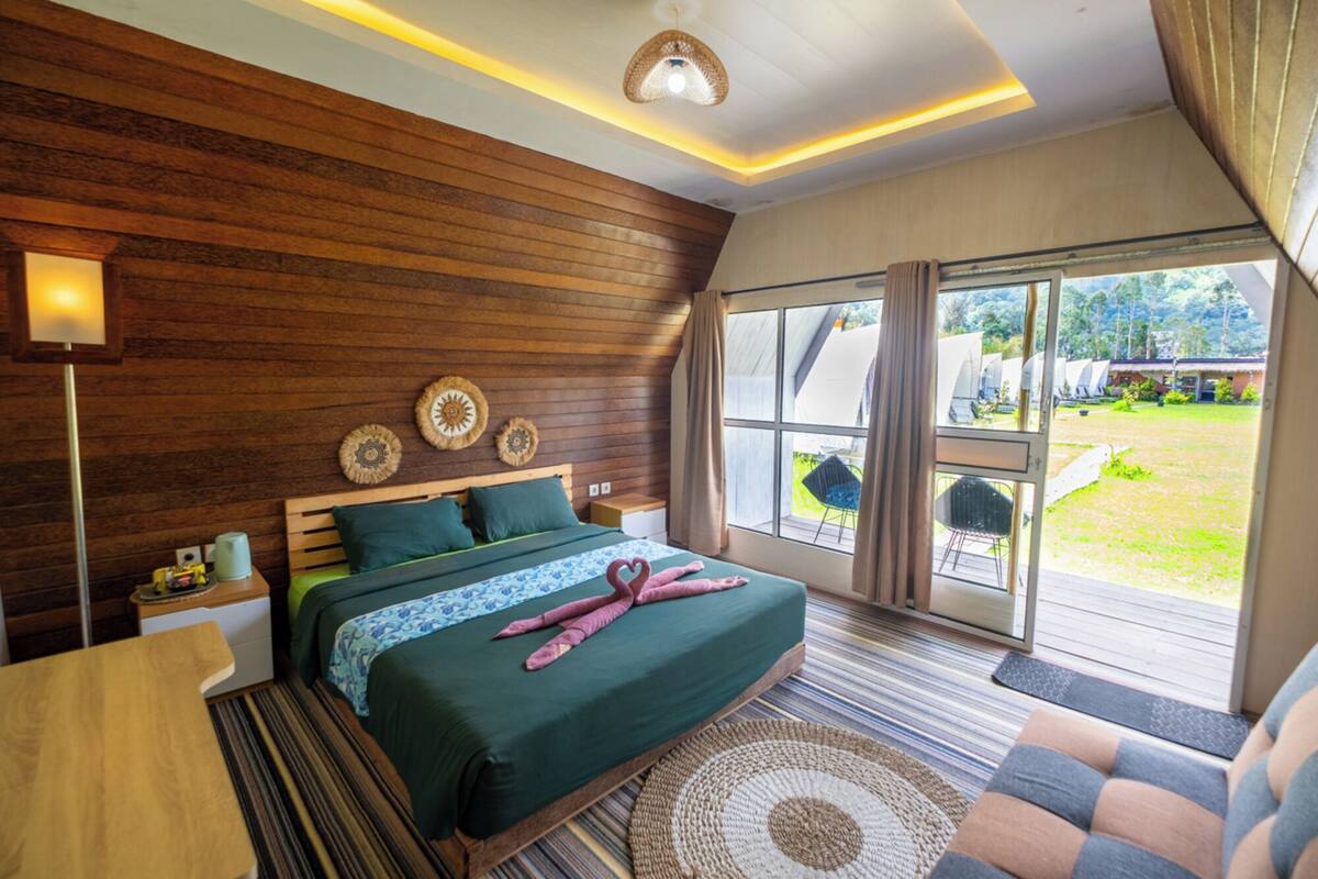 Une chambre avec un mur en bois, un lit avec une literie verte, une table de chevet, une lampe et une vue sur l’extérieur.