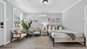 A modern bedroom with a bed, two chairs, a small table, a bedside lamp, and a large plant. - Sunny Pensacola Studio Near Beach & Downtown (Pensacola)