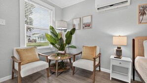 A room with a wooden floor, two white armchairs, a small round table with a plant, a nightstand with a lamp, and a bed with a headboard. - Sunny Pensacola Studio Near Beach & Downtown (Pensacola)