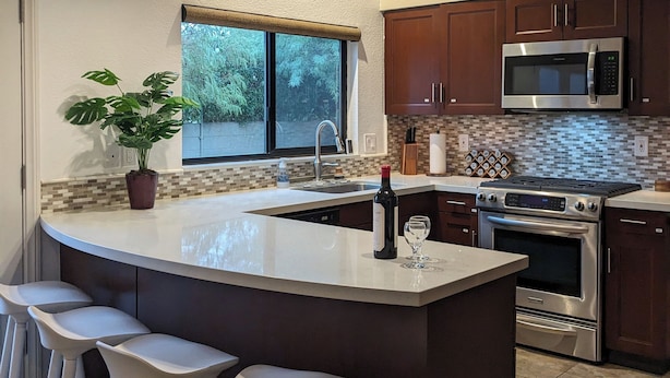 A modern kitchen with a large island, bar stools, a microwave, stove, and a potted plant.
