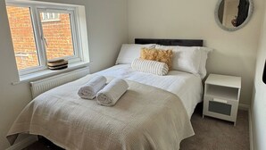 A neatly made bed with white linens and two rolled towels, a small bedside table, a round mirror, and a window with a view of a brick building. - Cottage - Sleeps 4 - Pets Ok - Garden - Parking (Middlewich)