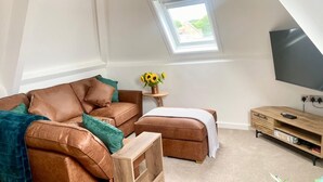 A modern loft bedroom with a sloped ceiling, a large window, a brown leather sofa, a wooden coffee table, a green armchair, and a TV on a wooden stand. - Central Tenby 3 Bed Apt Sleeps 6 to 8 Dog Friendly (Pembrokeshire)