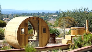 Uma estrutura em forma de barril de madeira, com porta e janelas, cercada por vegetação e vista para colinas ao longe.