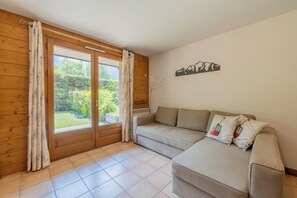 A room with a wooden wall, a sliding glass door to a garden, a beige sofa with pillows, and a wall-mounted artwork.