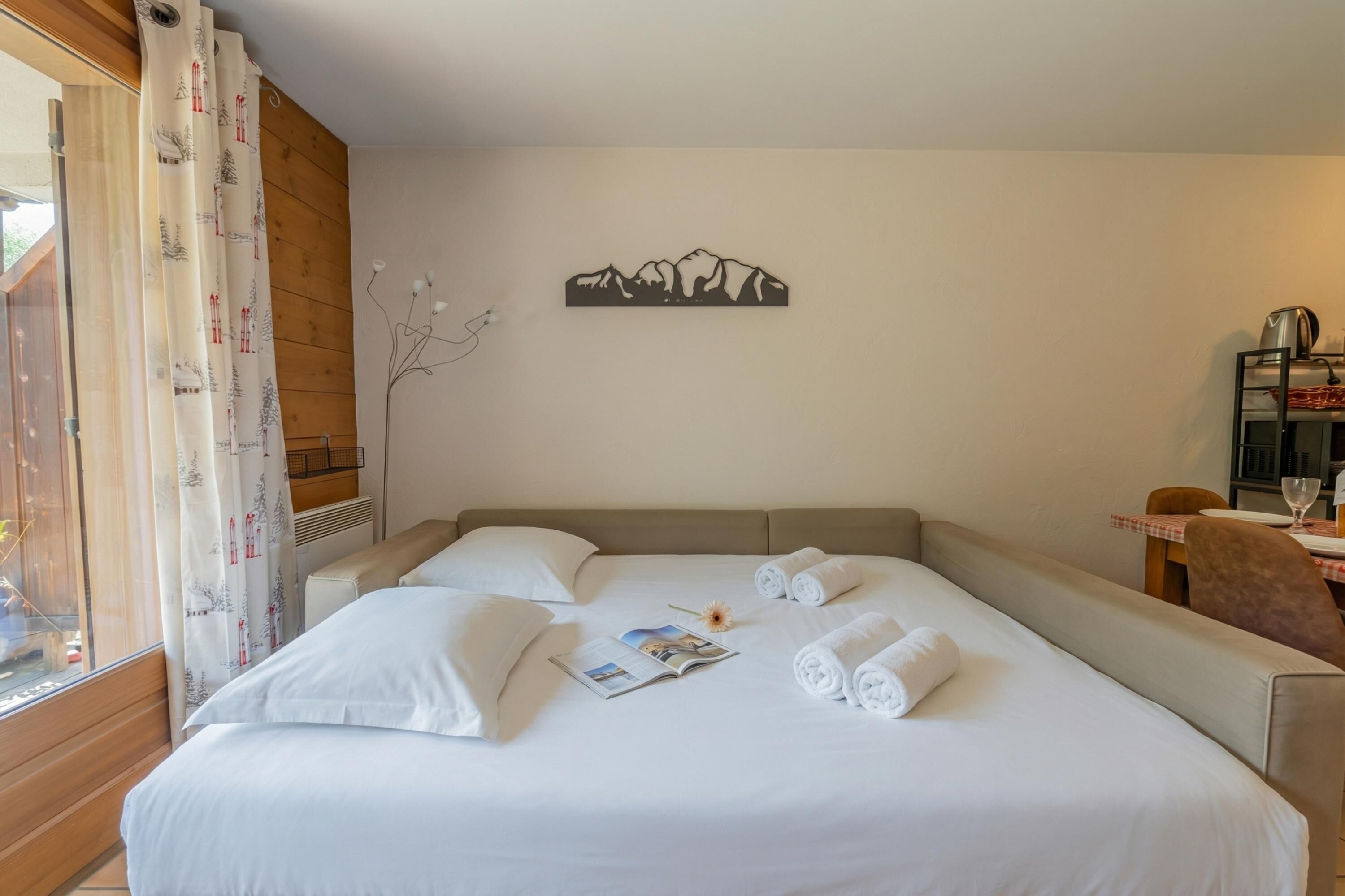 Un lit bien fait, avec du linge de lit blanc, une œuvre d’art représentant des montagnes accrochée au mur, et un coin repas avec une table et des chaises.