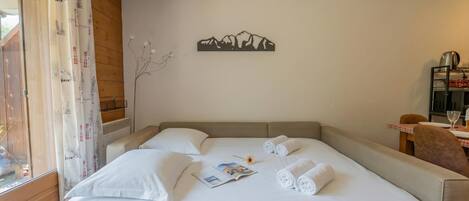 Un lit bien fait, avec du linge de lit blanc, une œuvre d’art représentant des montagnes accrochée au mur, et un coin repas avec une table et des chaises.