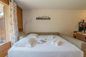 Ein ordentlich bezogenes Bett mit weißen Leinen, ein Wandbild von Bergen und ein Essbereich mit Tisch und Stühlen.