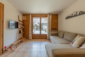 A cozy living room with a sofa, a wooden ladder, a TV, and a view of a snowy landscape through a glass door. - Apt Les Capucins Quiet Chamonix Base w/ Private Garden, Chamonix, France (Chamonix)