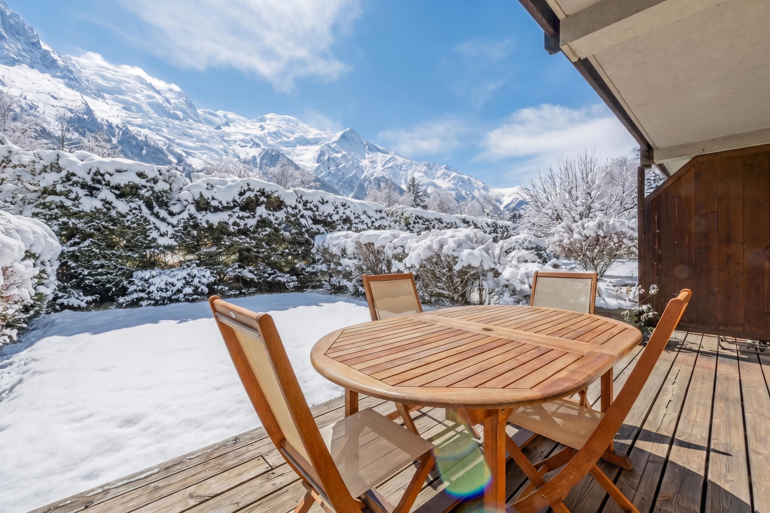 A wooden deck with a round table and chairs, overlooking a snowy mountain landscape.