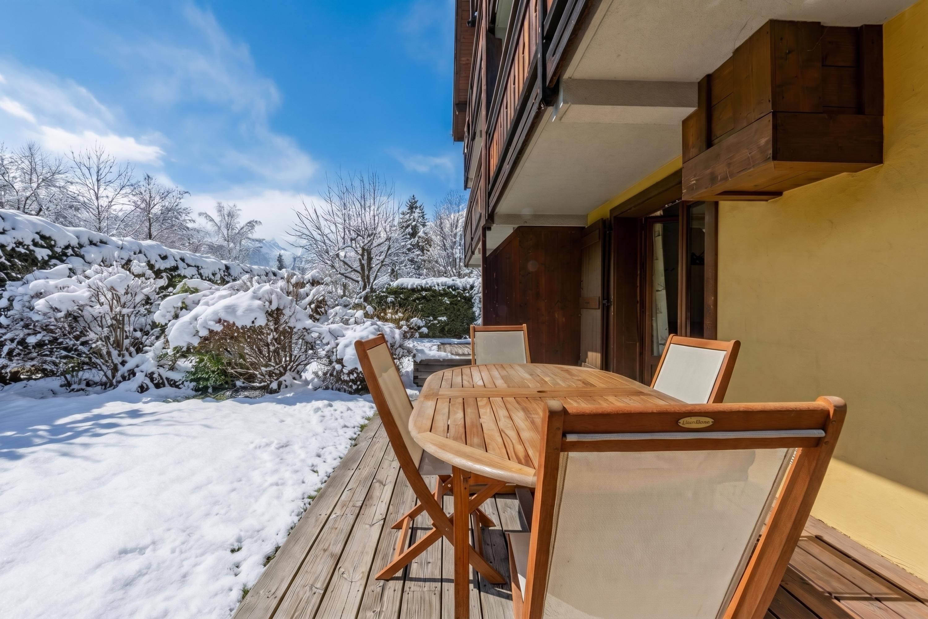 A wooden deck with a table and chairs, a building with a balcony, and a snowy landscape.