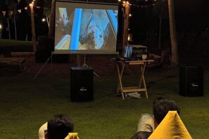 Un écran de projection diffusant un film, un haut-parleur et un support en bois, le tout installé dans un cadre extérieur.