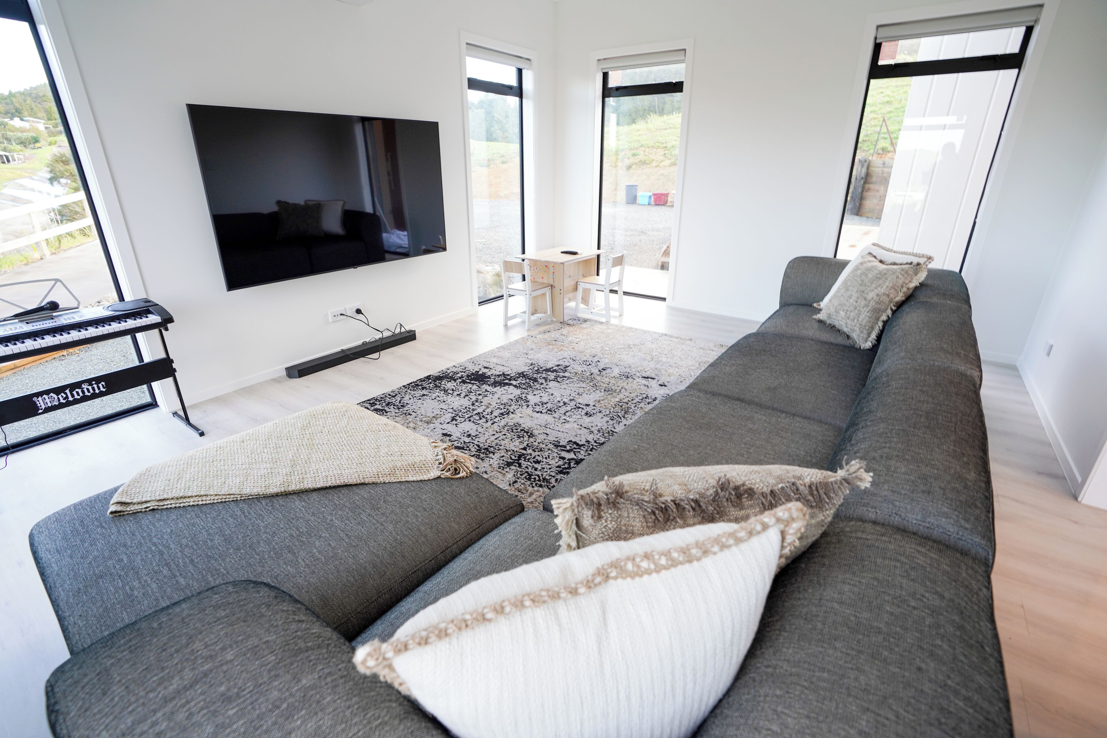 A modern living room with a grey sofa, a flat-screen TV, a keyboard, and a large window offering a view of the outdoors.
