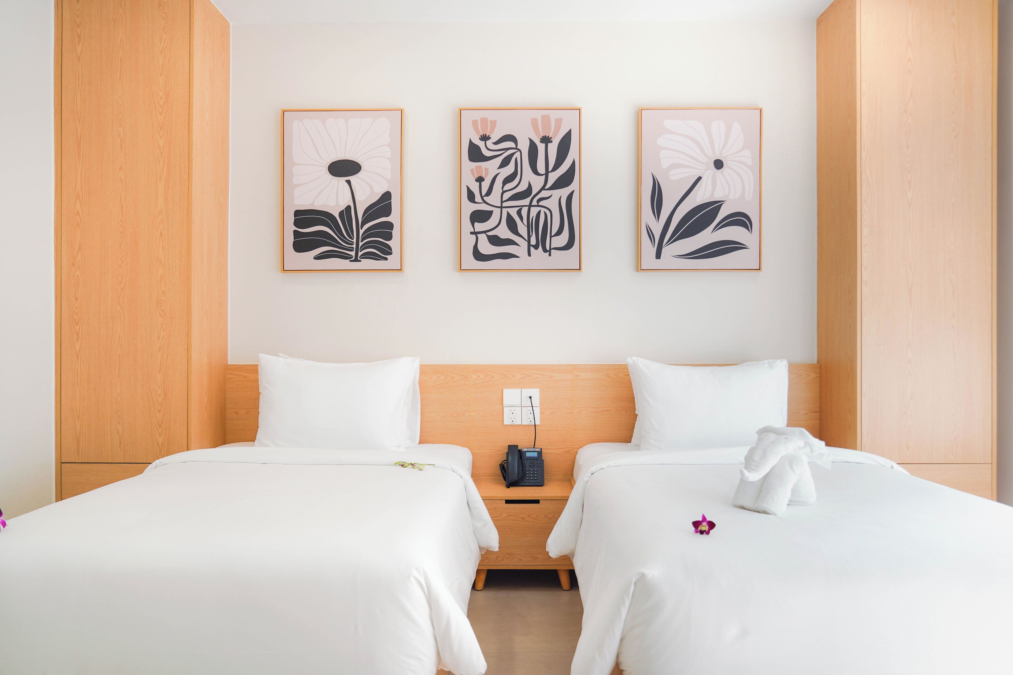 A hotel room with two beds, a wooden headboard, a nightstand, and three framed floral artworks on the wall.