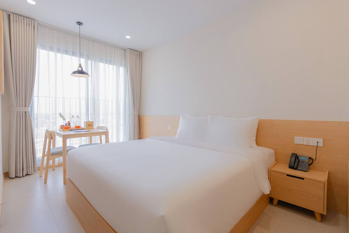 A hotel room with a large bed, a bedside table with a phone, a desk with a chair, and a window with curtains.