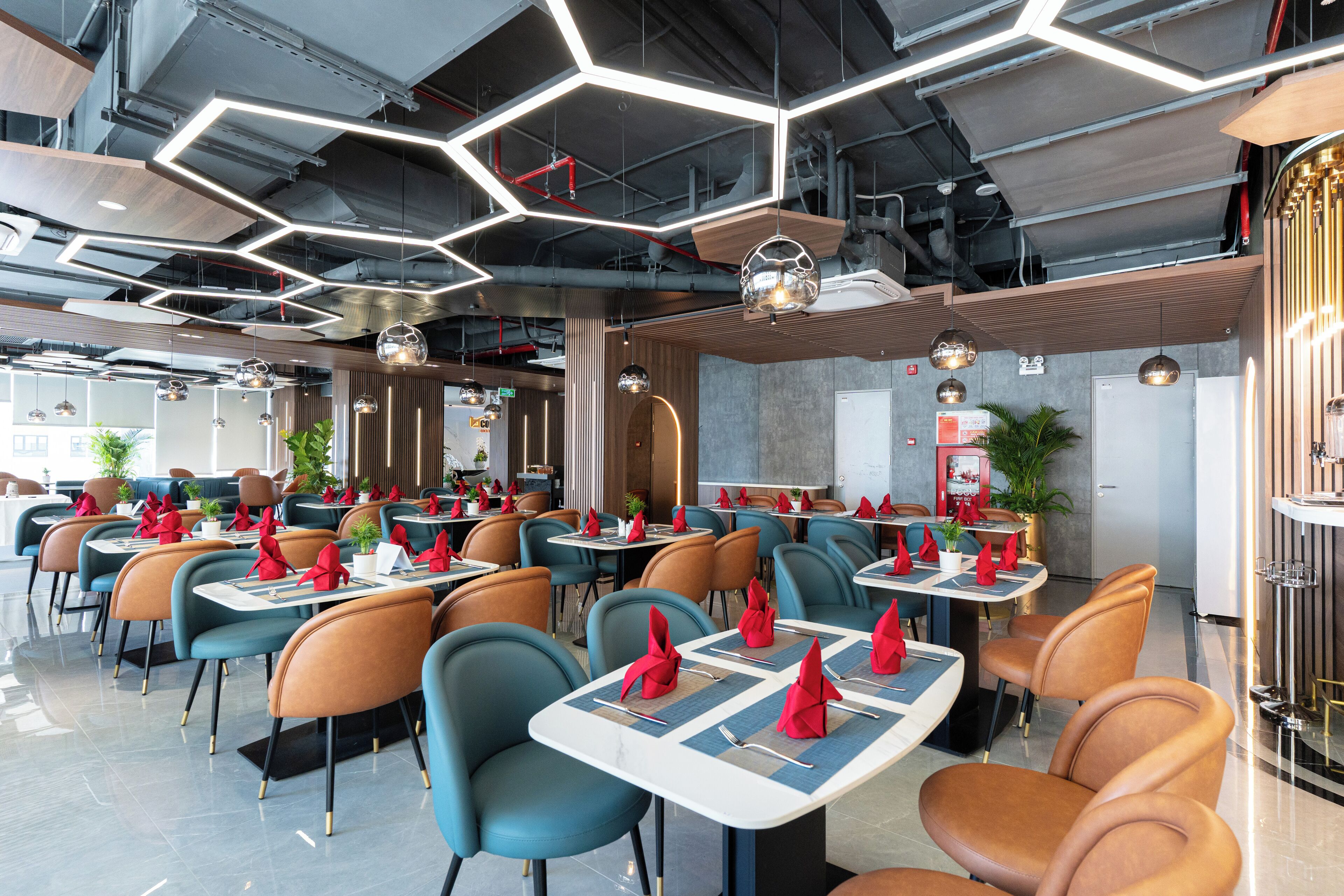 A modern restaurant with hexagonal ceiling lights, blue and orange chairs, and neatly set tables with red napkins.