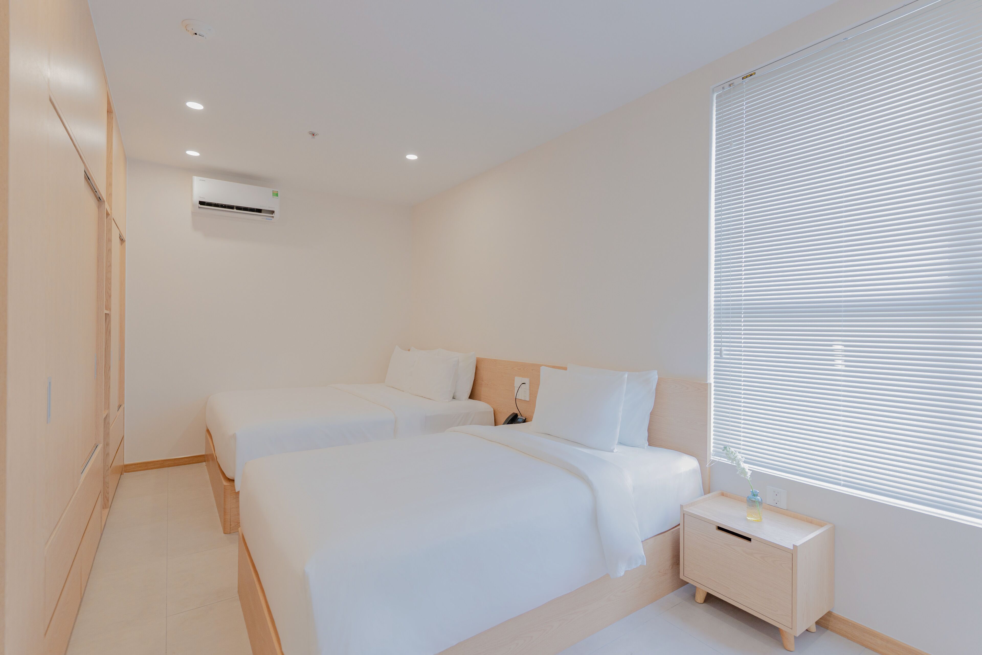 A modern hotel room with two beds, a bedside table, a nightstand, and a window with blinds.