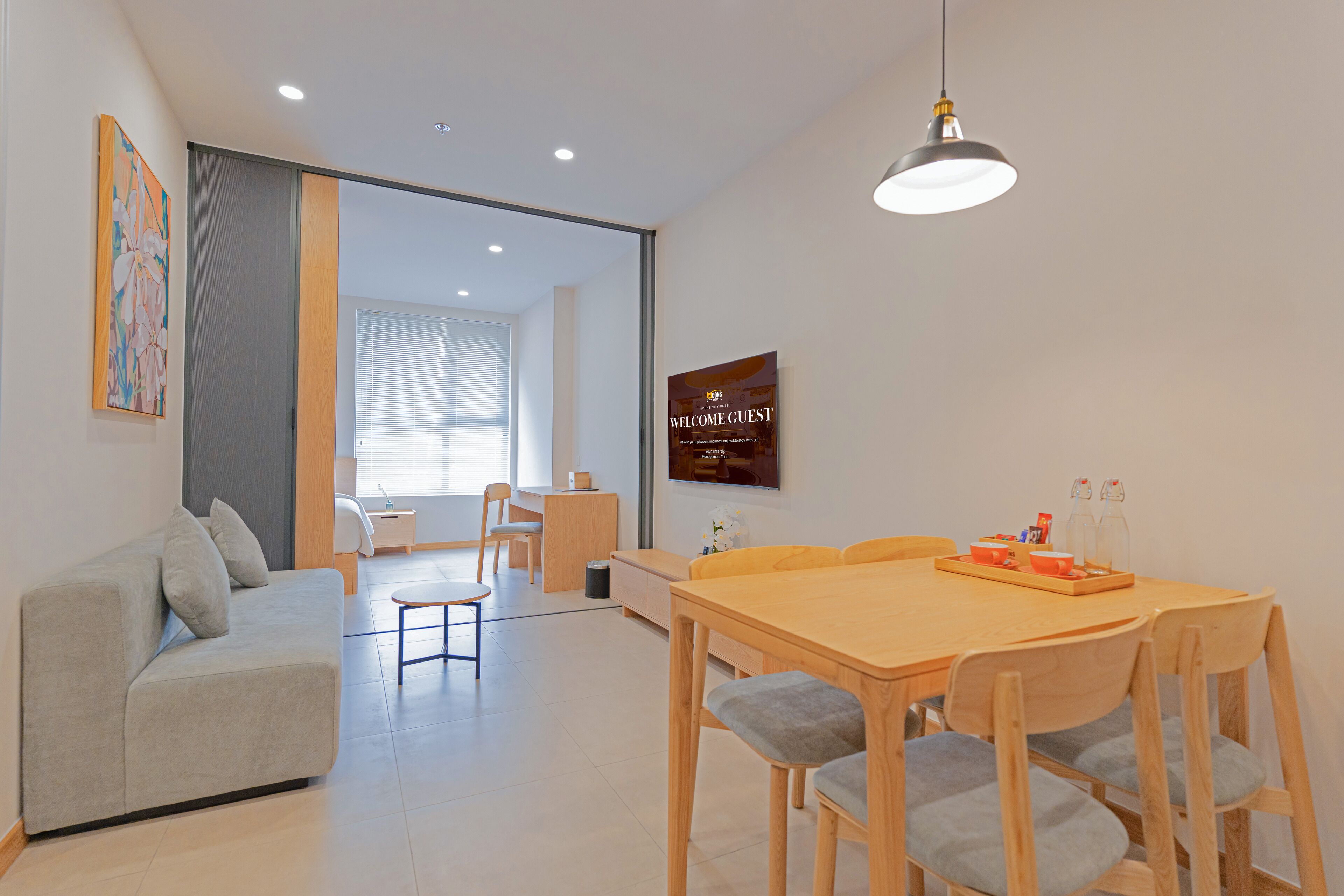 A modern living room with a sofa, dining table, and chairs. There is a television mounted on the wall and a window with blinds.