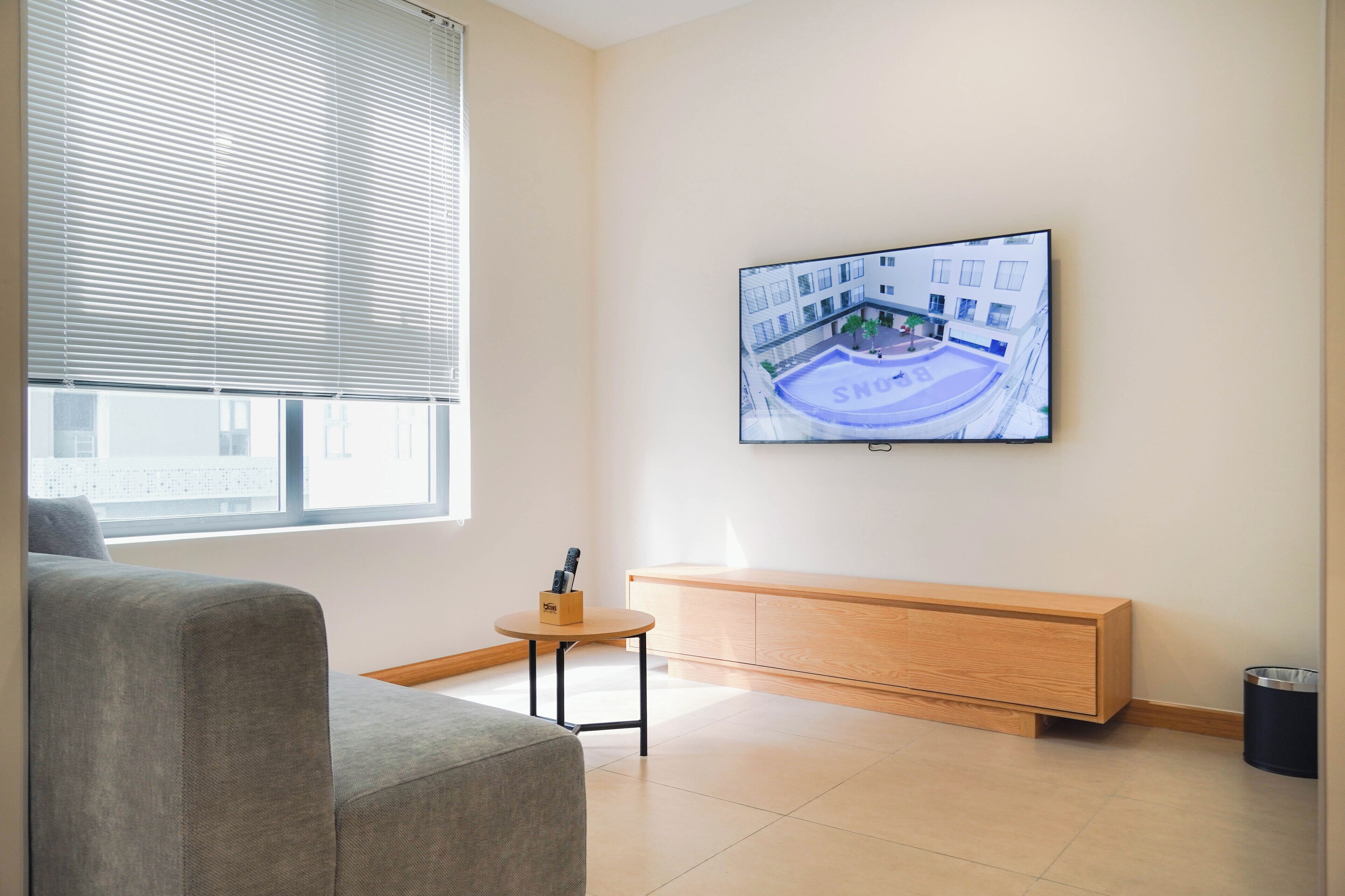 A modern living room with a grey sofa, a wooden bench, a small round table with a pen holder, and a TV mounted on the wall displaying an aerial view of a building.