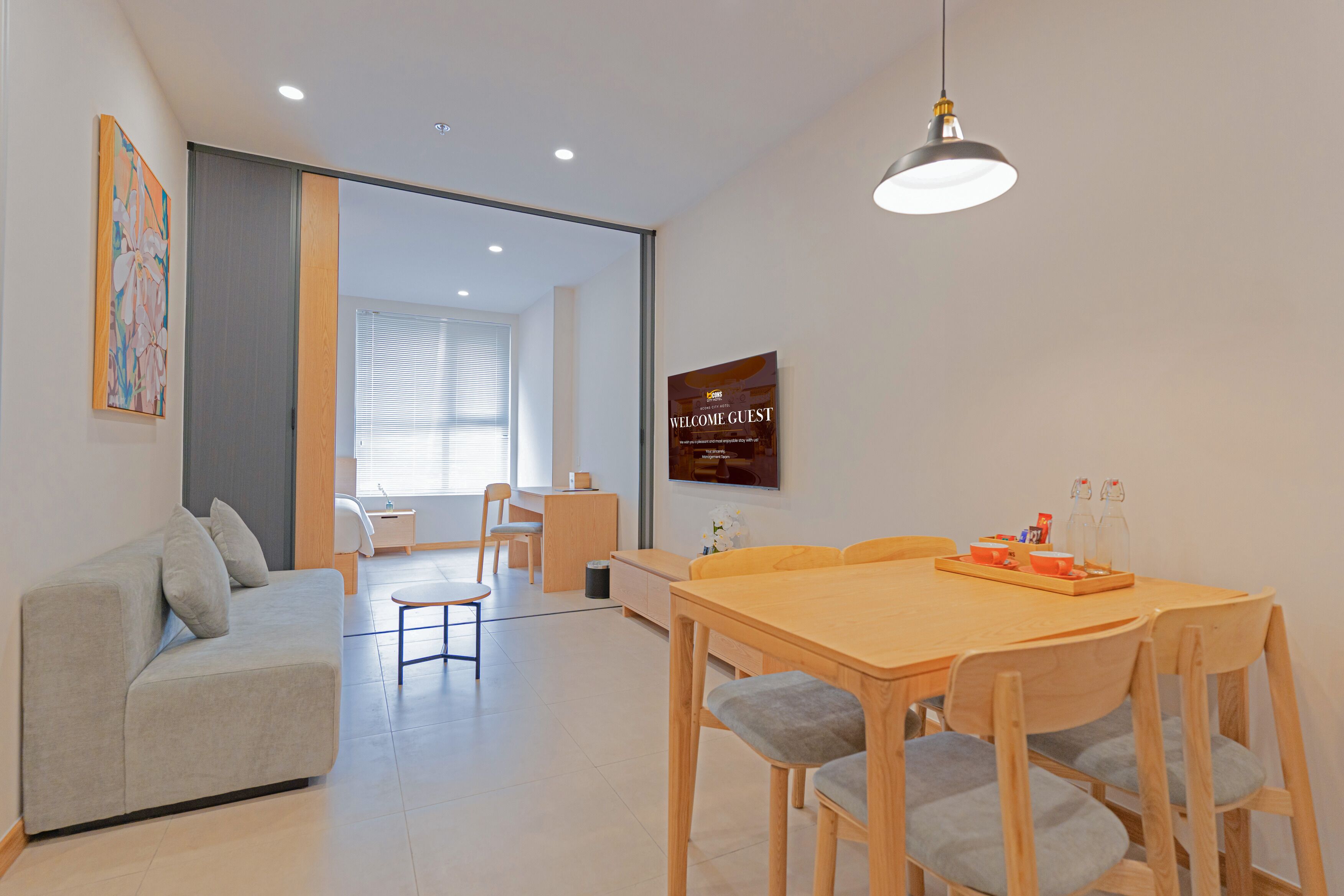 A modern living room with a sofa, dining table, and chairs. There is a television mounted on the wall and a sign that reads "WELCOME GUEST".